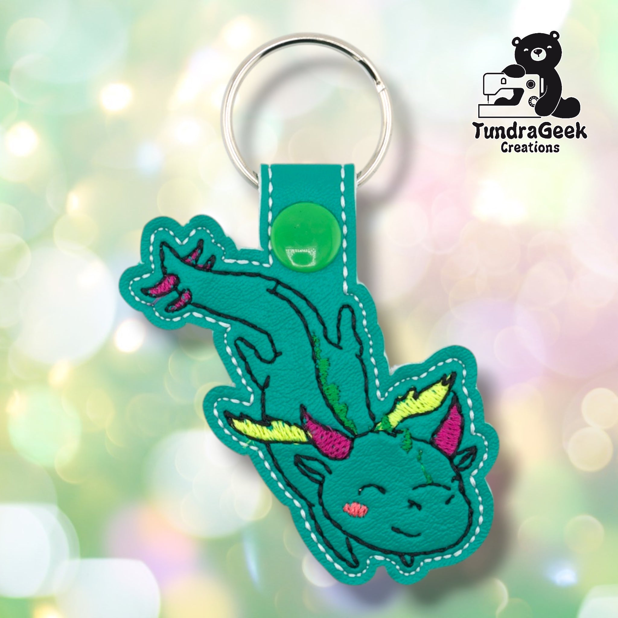 Teal faux leather keychain shaped like a dragon in a playful tumbling pose. Black embroidery details the body; wings are embroidered in yellow-green and pink, with pink horns and green spines along the back. The dragon's chubby round head features a smiling expression and a rosy pink cheek. Green plastic snap closure and silver key ring at top. TundraGeek Creations logo in upper right corner.