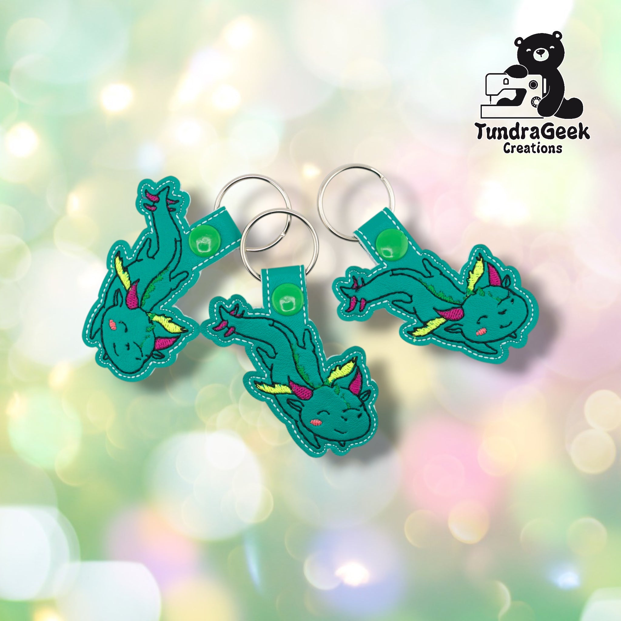 Three whimsical dragon keychains arranged together on a pastel bokeh background, showing the design from varied angles. All three feature matching teal base, multicolor embroidery, and green snap closures.
