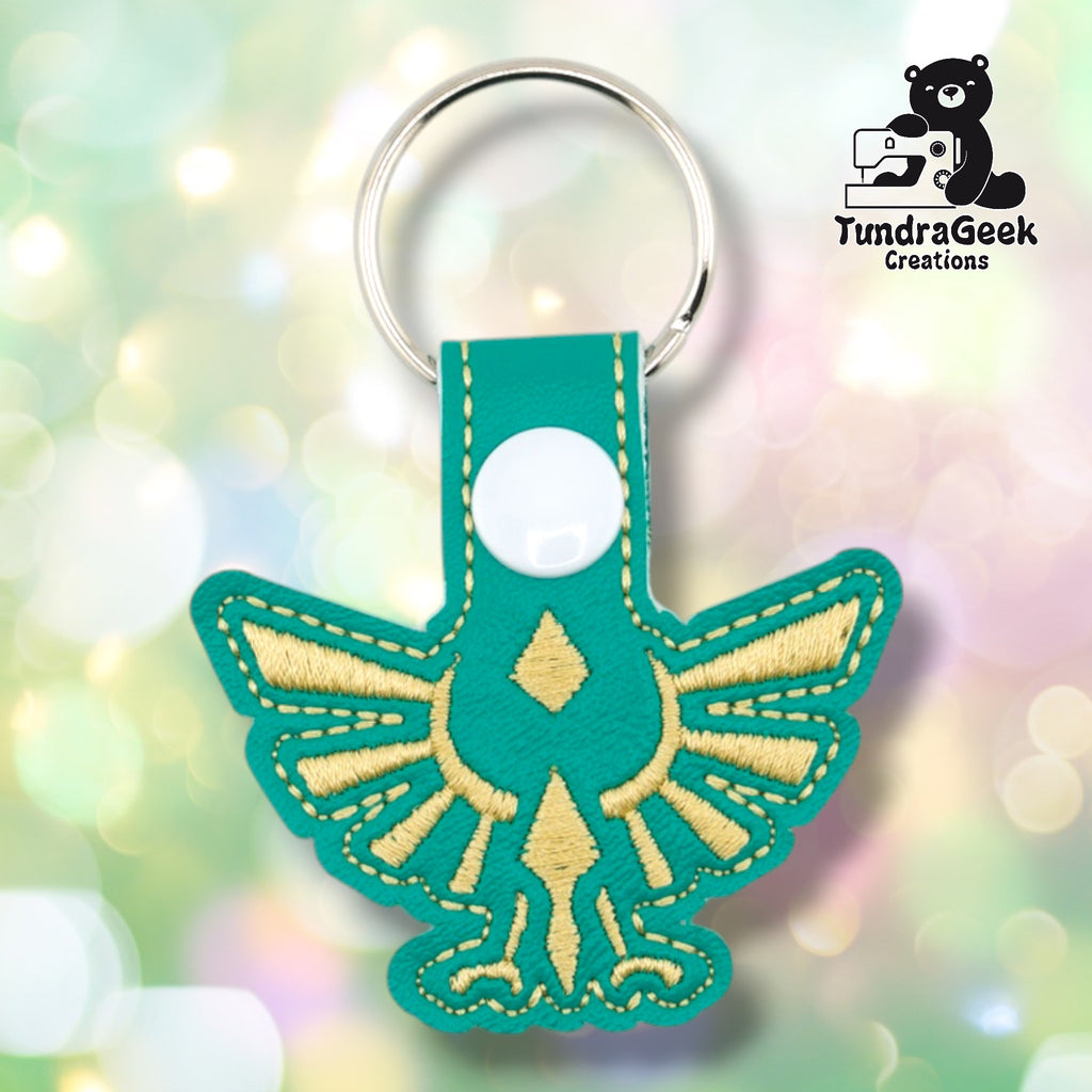 Teal faux leather keychain shaped like a winged heraldic crest from a classic fantasy video game. Gold embroidery details the spread wings, central diamond shape, and downward-pointing element of the iconic design. White plastic snap closure and silver key ring at top. TundraGeek Creations logo in upper right corner.