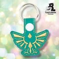 Teal faux leather keychain shaped like a winged heraldic crest from a classic fantasy video game. Gold embroidery details the spread wings, central diamond shape, and downward-pointing element of the iconic design. White plastic snap closure and silver key ring at top. TundraGeek Creations logo in upper right corner.