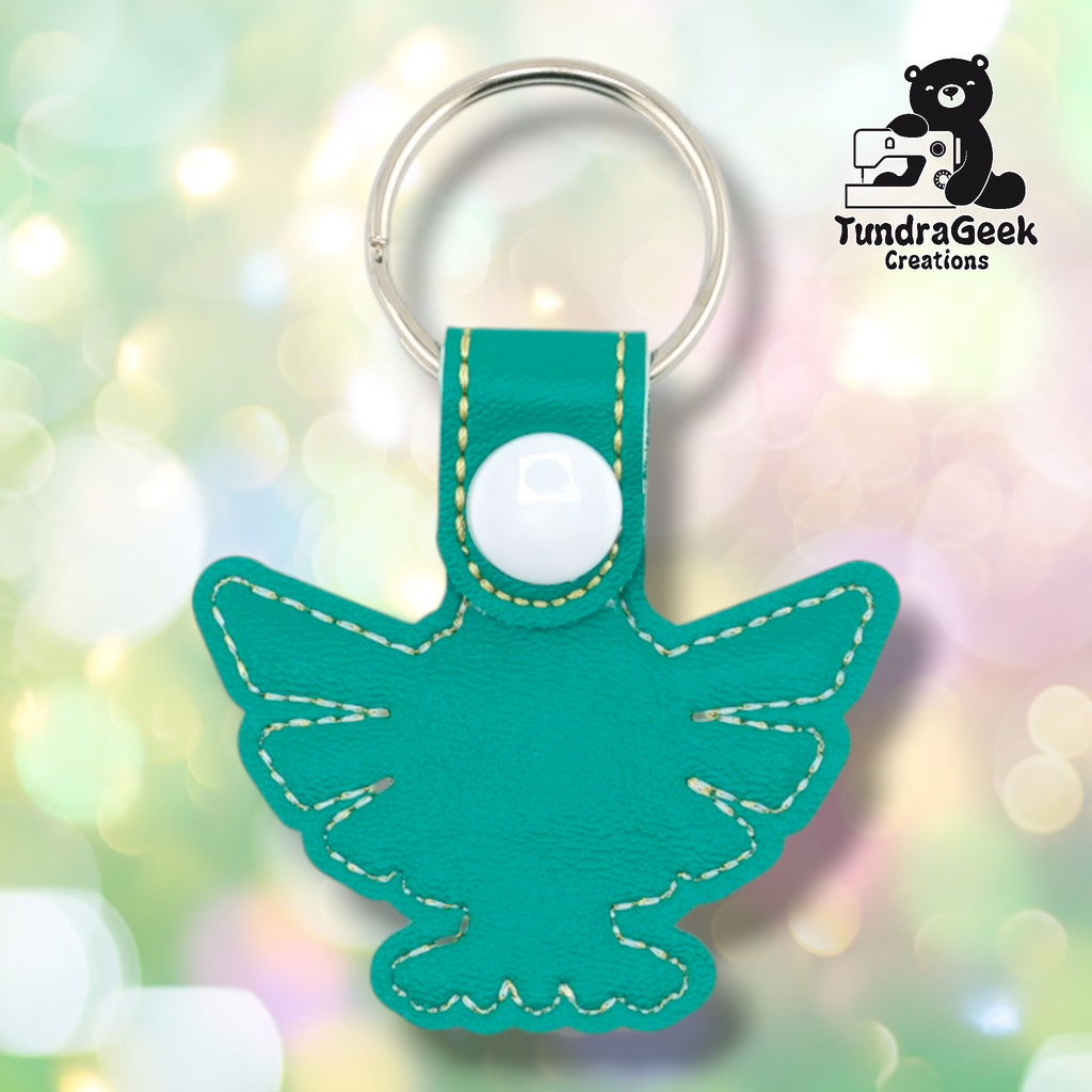 Plain back of the Hylian crest keychain showing teal faux leather and gold outline stitching following the winged crest silhouette. White snap closure at top. No embroidery on reverse.