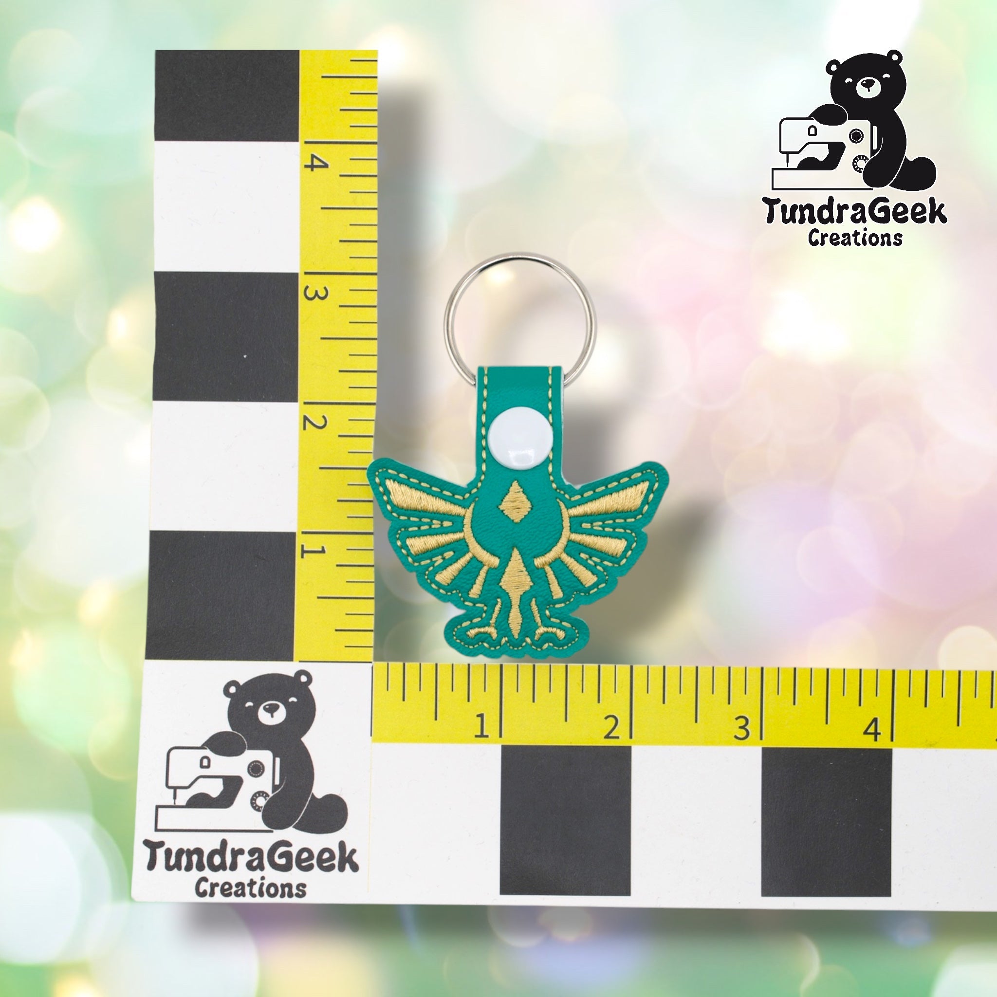 Hylian crest keychain positioned against a TundraGeek Creations branded ruler card showing yellow tape measure. Keychain body measures approximately 2.5 inches wide by 1.5 inches tall, not including hardware.