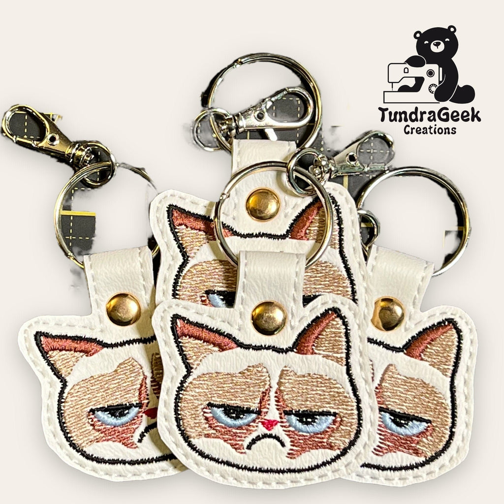 Three keychains with embroidered grumpy cat faces on white material, displayed overlapping. TundraGeek Creations logo with a bear is in the top right corner.