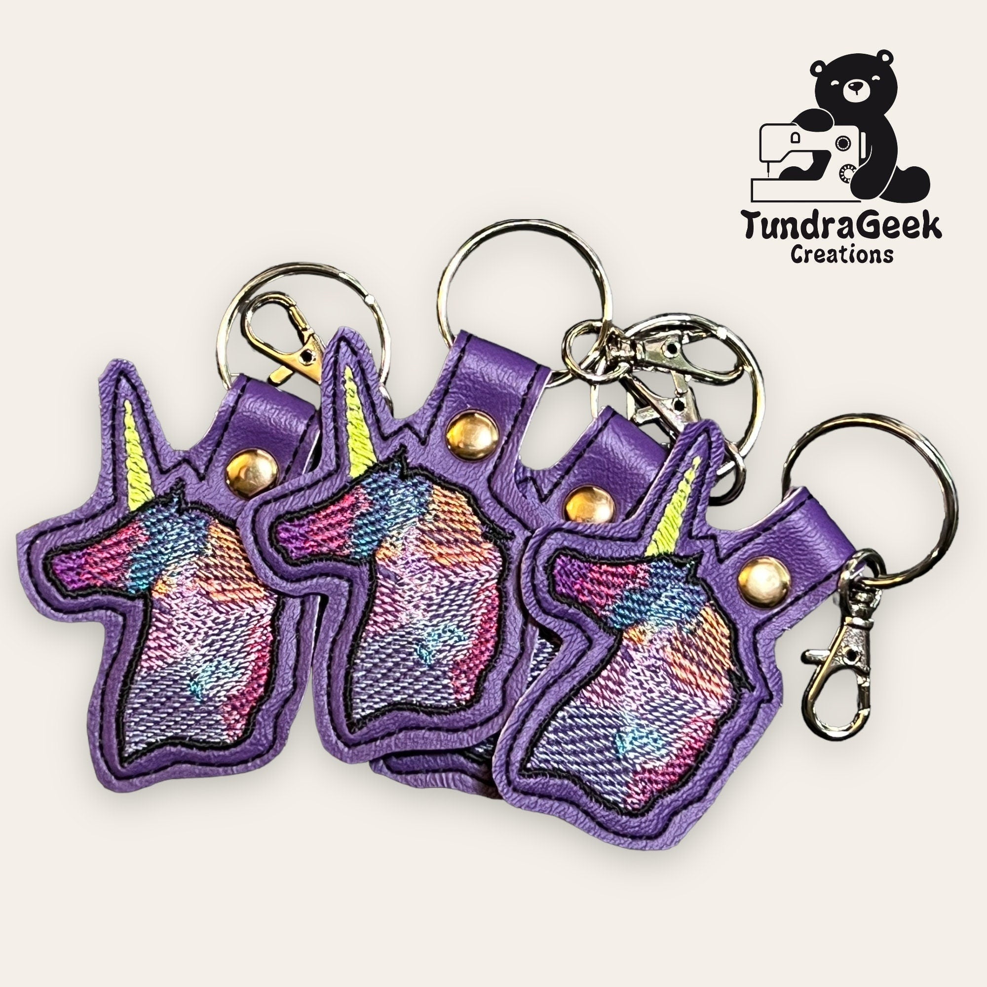 Four purple unicorn-shaped keychains with multicolored embroidered designs are displayed. The TundraGeek Creations logo with a bear and text appears in the upper right corner.
