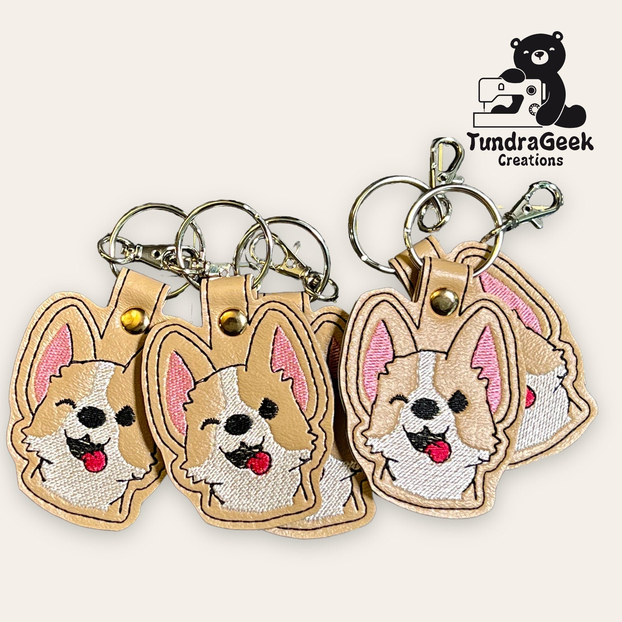 Five embroidered keychains featuring cartoon-style corgi faces with various expressions, displayed together; TundraGeek Creations logo in the corner.
