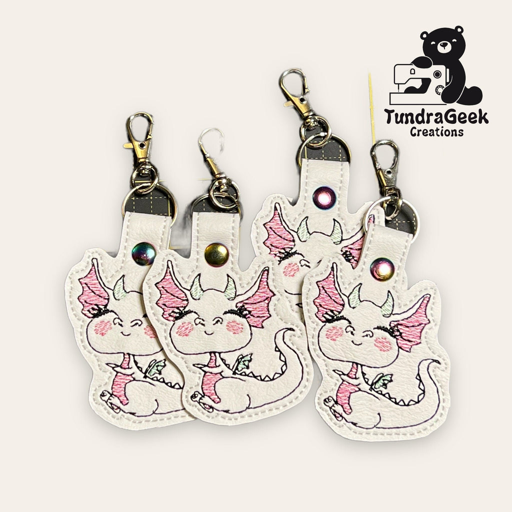 Four white keychain holders featuring a cute, embroidered dragon with pink wings and cheeks, displayed with metal clasps. TundraGeek Creations logo appears in the top right corner.