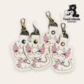 Four white keychain holders featuring a cute, embroidered dragon with pink wings and cheeks, displayed with metal clasps. TundraGeek Creations logo appears in the top right corner.
