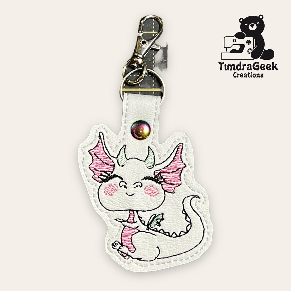 White faux leather keychain featuring a smiling dragon with pink details, metal clasp, and the TundraGeek Creations logo in the top right corner.