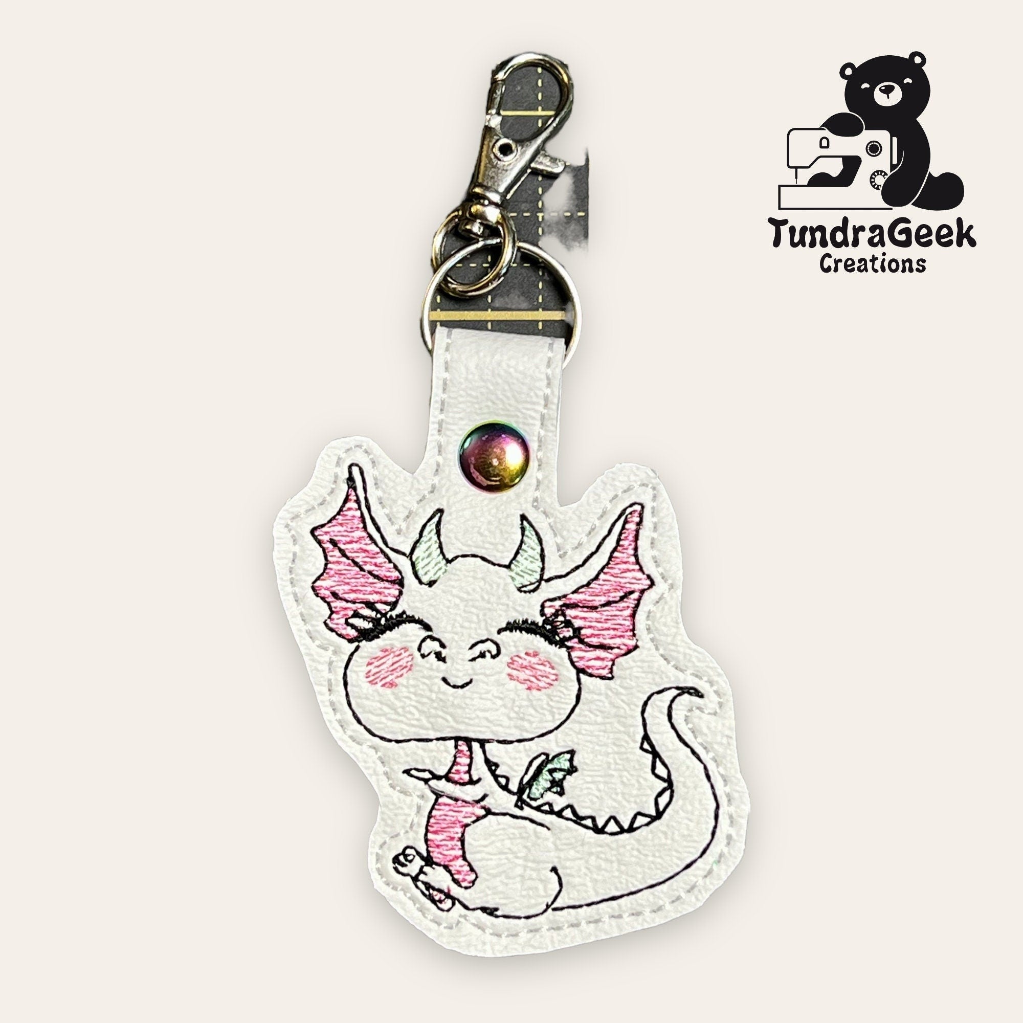 White faux leather keychain featuring a smiling dragon with pink details, metal clasp, and the TundraGeek Creations logo in the top right corner.