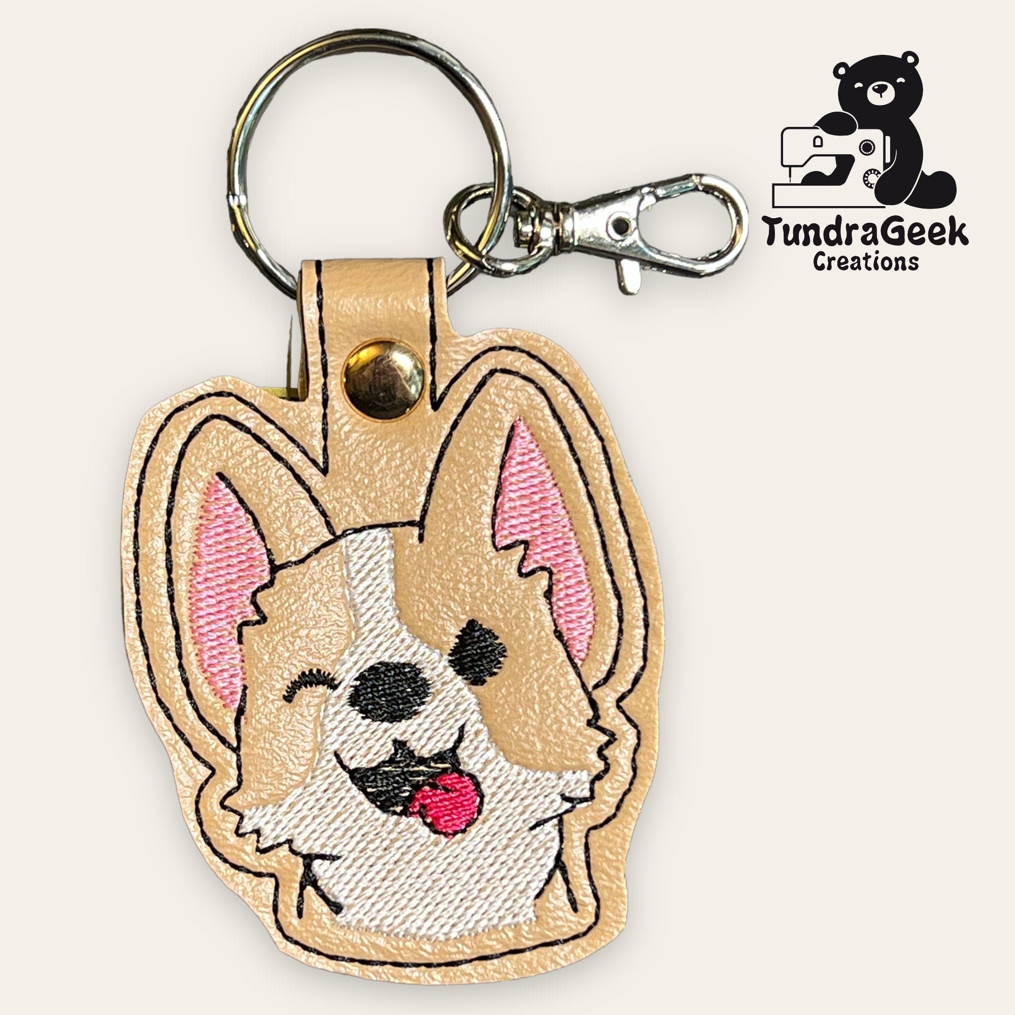 A keychain with an embroidered cartoon corgi face, attached to a metal key ring and clasp, with the TundraGeek Creations logo in the corner.