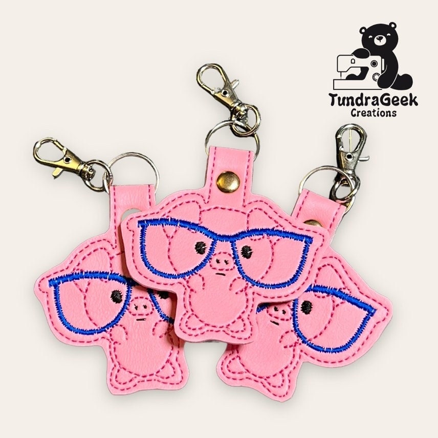 Three pink pig-shaped keychains with blue embroidered glasses are arranged together. The TundraGeek Creations logo is in the top right corner.