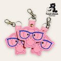 Three pink pig-shaped keychains with blue embroidered glasses are arranged together. The TundraGeek Creations logo is in the top right corner.