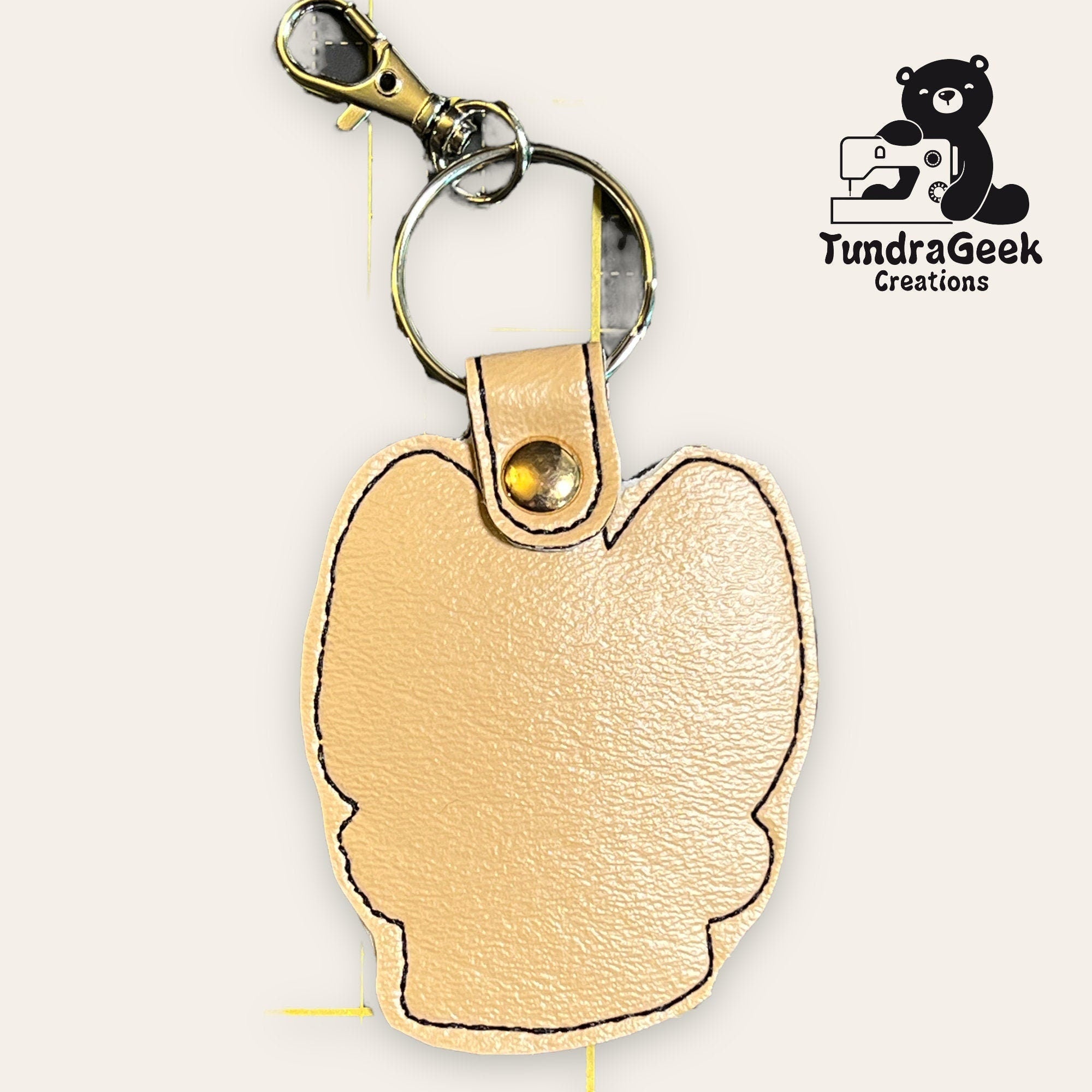 Tan leather keychain shaped like an animal head, attached to a metal key ring and clasp. TundraGeek Creations logo with a bear and sewing machine is in the top right corner.