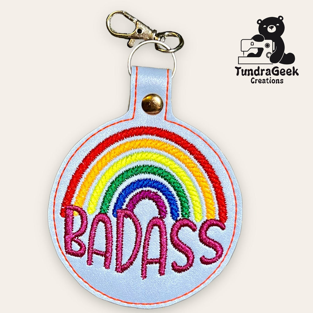 A round keychain with a clasp, featuring an embroidered rainbow and the word BADASS in pink thread. TundraGeek Creations logo is in the top right corner.