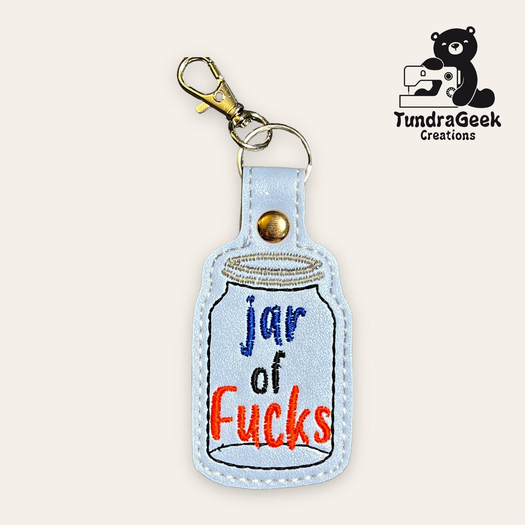 Keychain shaped like a jar with the words jar of Fucks embroidered on it, and a metal clasp attached. TundraGeek Creations logo in the top right corner.