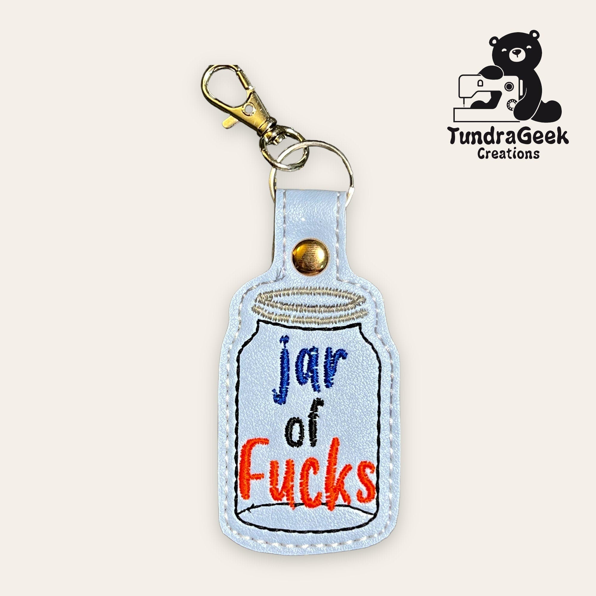 Keychain shaped like a jar with the words jar of Fucks embroidered on it, and a metal clasp attached. TundraGeek Creations logo in the top right corner.