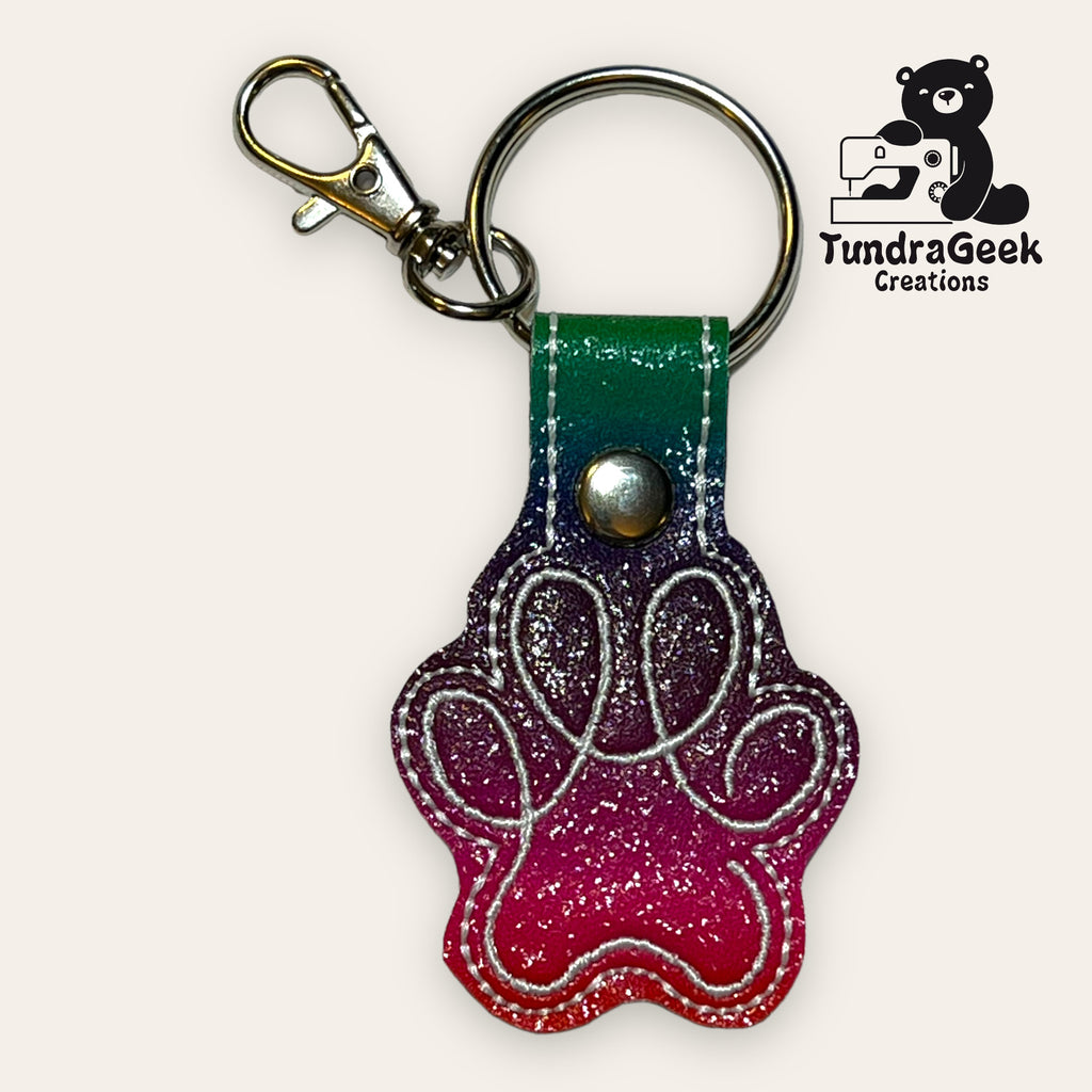 Paw-shaped keychain with a pink-to-green gradient, metal clasp, and keyring. TundraGeek Creations logo with a bear and sewing machine in the top right corner.