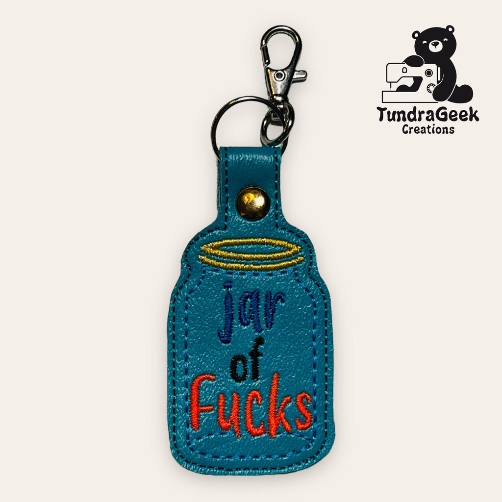 A blue embroidered keychain shaped like a jar with the words jar of fucks in colorful letters, and a metal clasp attached at the top. TundraGeek Creations logo is in the corner.