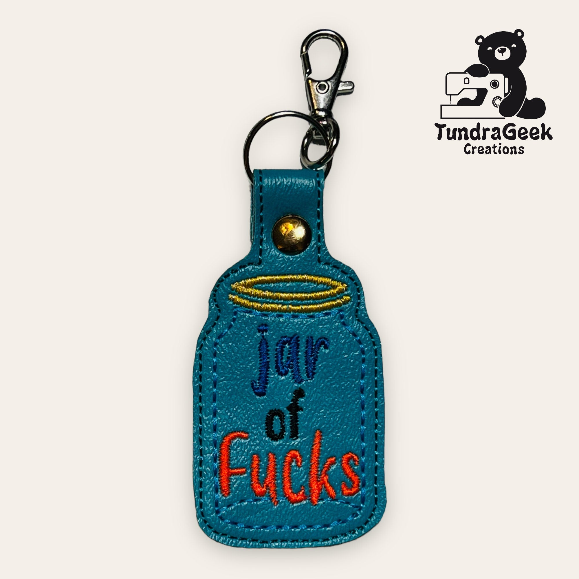 A blue keychain shaped like a jar with the words jar of fucks embroidered in colorful letters, displayed with a metal clip and ring. TundraGeek Creations logo is in the top right corner.