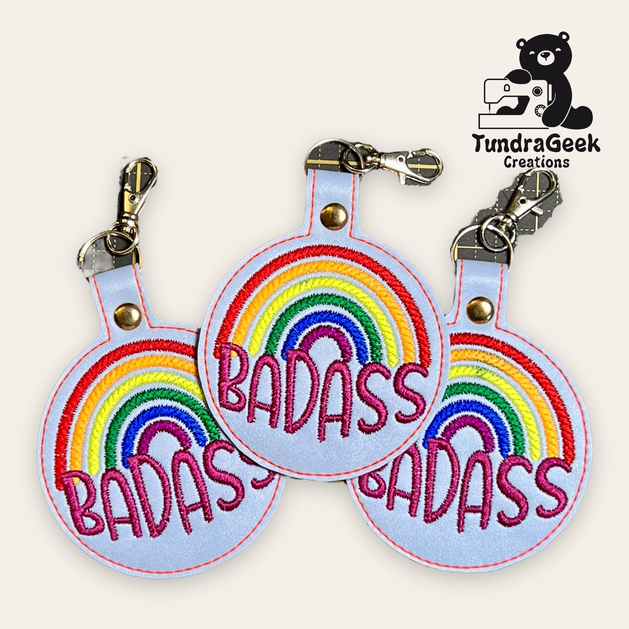 Three round keychains with rainbow embroidery and the word BADASS in bold letters, displayed with TundraGeek Creations logo in the corner.