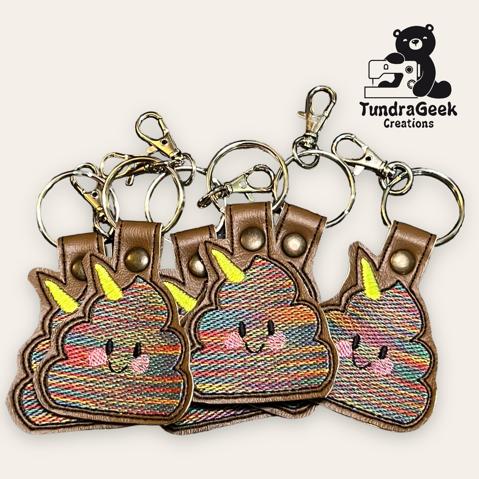 Four embroidered keychains shaped like smiling rainbow poop with yellow horns are arranged together. The TundraGeek Creations logo with a bear is in the top right corner.