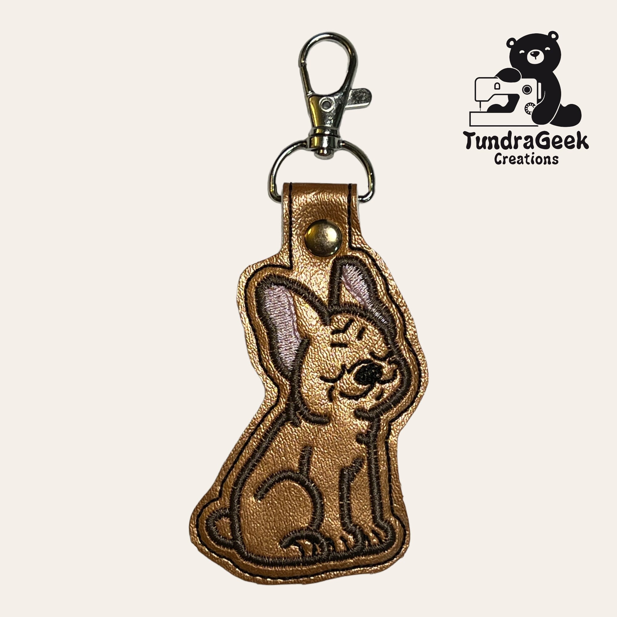 Brown faux leather keychain shaped like a sitting Chihuahua dog, with embroidered details and a metal clasp. TundraGeek Creations logo is visible in the top right corner.