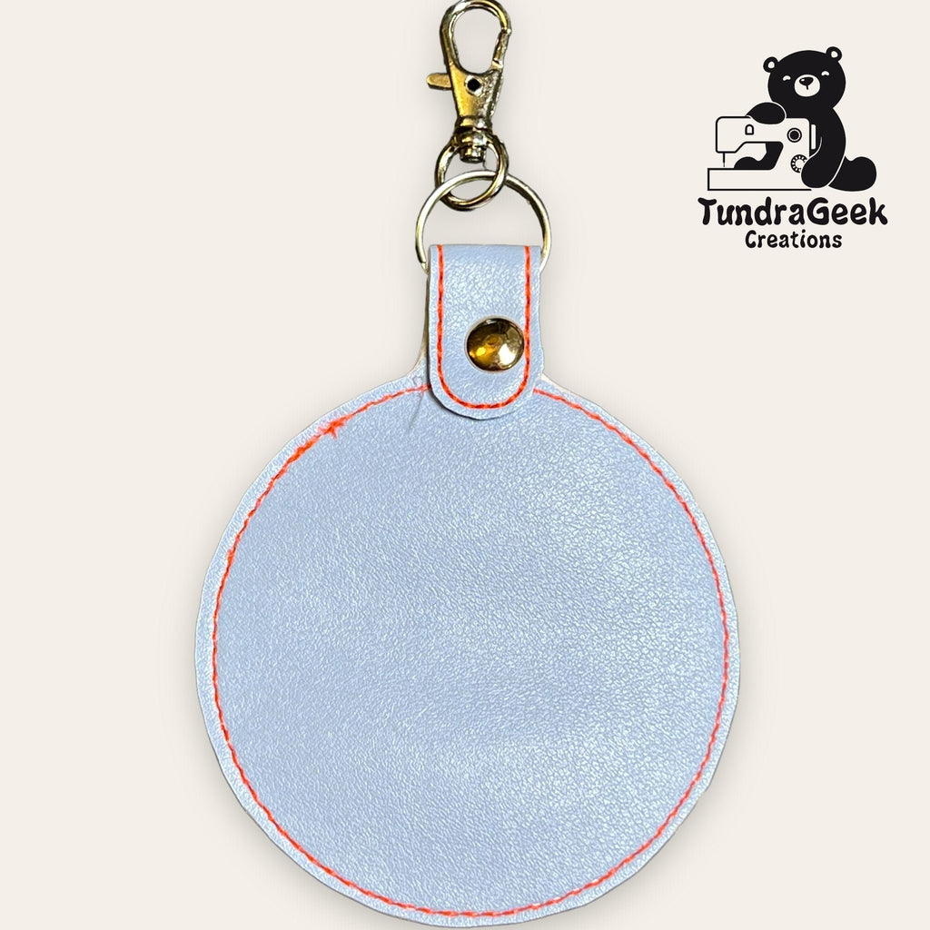 Round light gray leather keychain with red stitching, metal clasp, and rivet. TundraGeek Creations logo with bear and sewing machine in the top right corner.