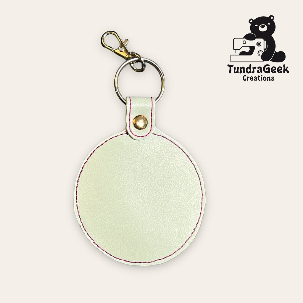 Round white faux leather keychain with pink stitching, metal rivet, keyring, and clasp. TundraGeek Creations logo featuring a bear with a sewing machine in the top right corner.