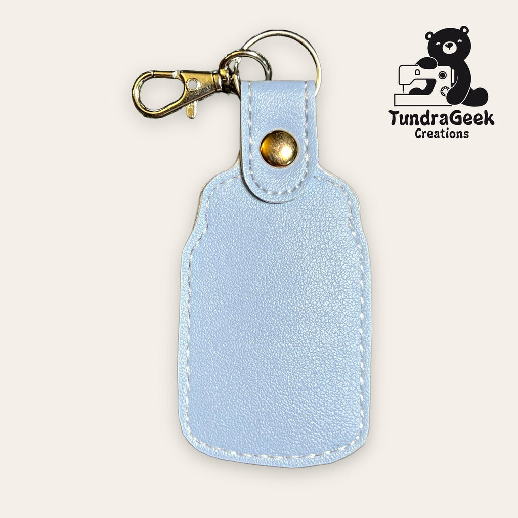 Light blue faux leather keychain holder with gold snap button, metal keyring, and lobster clasp. TundraGeek Creations logo with a bear and sewing machine in the top right corner.