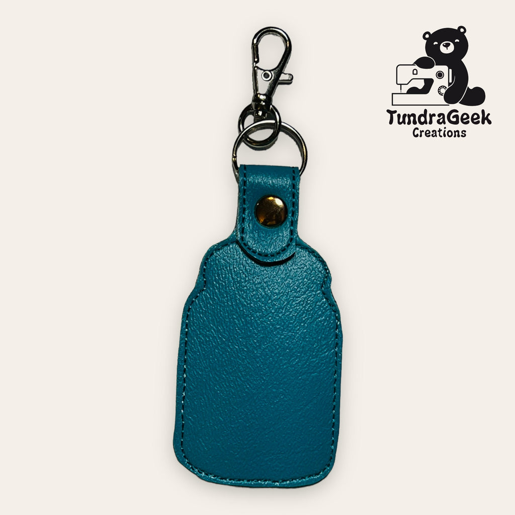 Teal leather keychain holder with brass snap and silver clip, displayed against a plain background with the TundraGeek Creations logo in the top right corner.