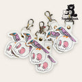 A group of embroidered keychains featuring a purple bird perched on a white skull with pink accents, displayed with metal clips.