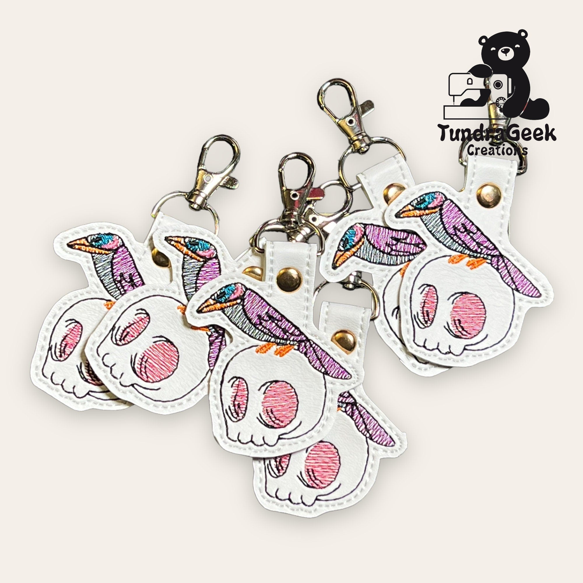 A group of embroidered keychains featuring a purple bird perched on a white skull with pink accents, displayed with metal clips.