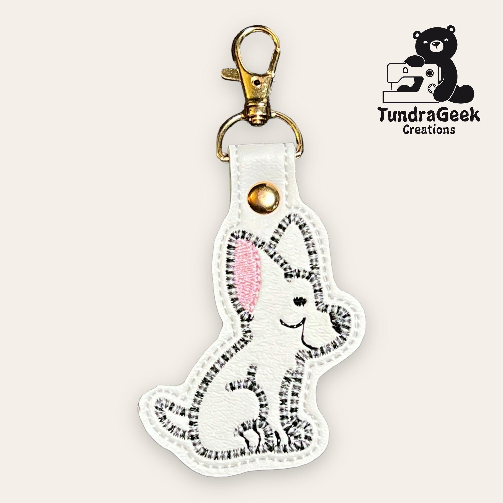 A white keychain shaped like a smiling dog with pink ears, gold hardware, and TundraGeek Creations logo featuring a bear and sewing machine in the corner.