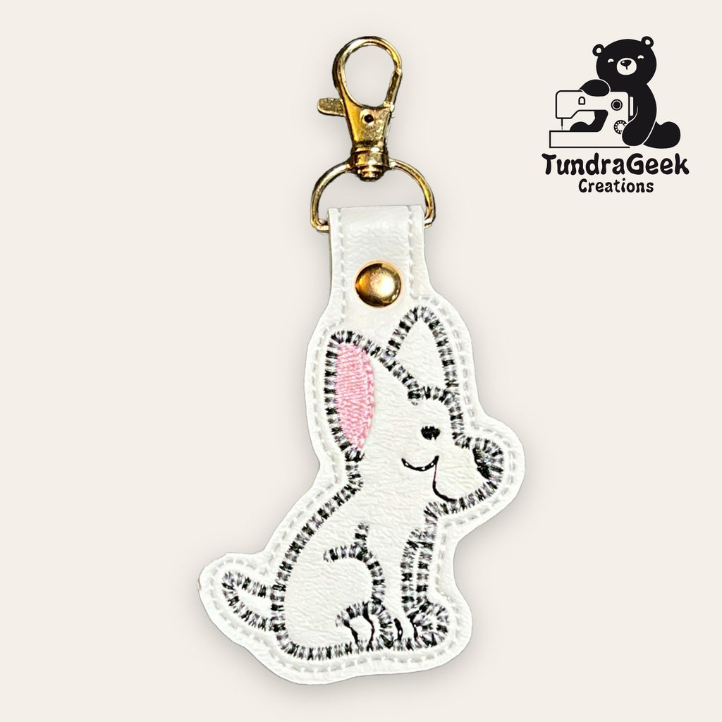 A white faux leather keychain featuring an embroidered cartoon dog with pink ears, next to the TundraGeek Creations logo.