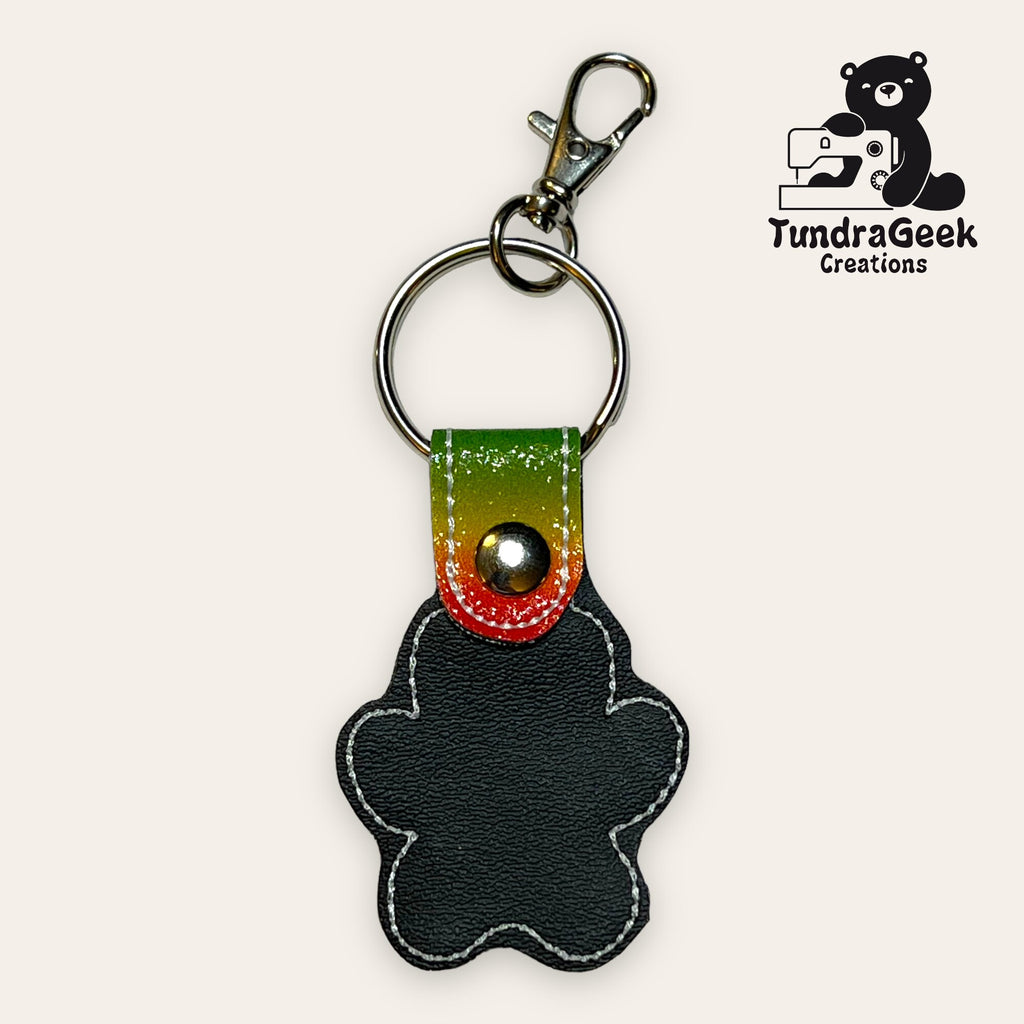 Keychain shaped like a gingerbread person with black faux leather, white stitching, and a multicolored top; includes metal key ring and clasp. TundraGeek Creations logo in the corner.