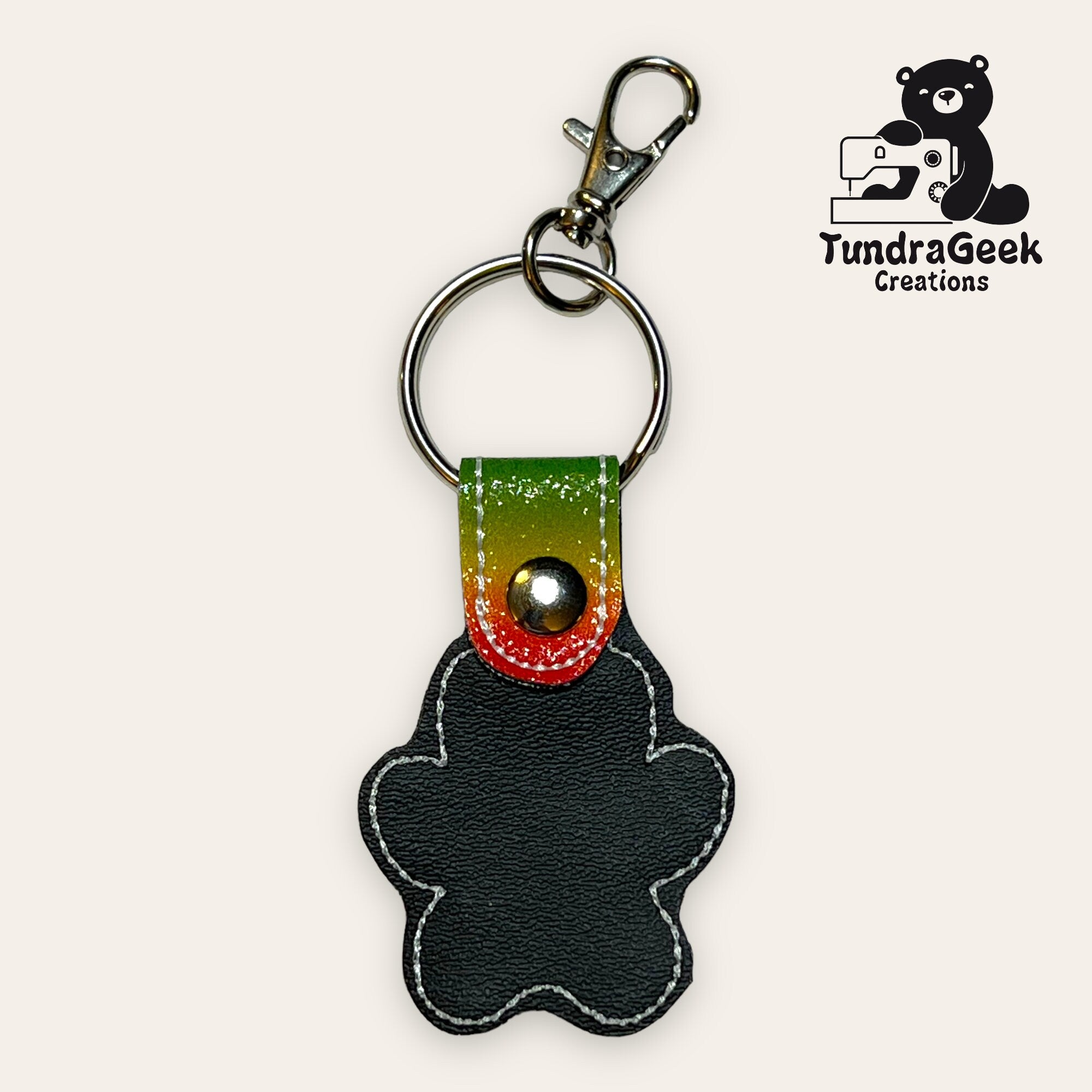 Keychain shaped like a gingerbread person with black faux leather, white stitching, and a multicolored top; includes metal key ring and clasp. TundraGeek Creations logo in the corner.