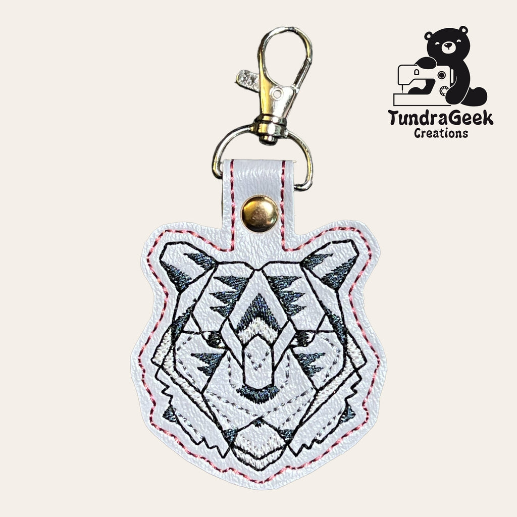 White leather keychain shaped like a tigers head with black geometric embroidery. Gold clasp attached. TundraGeek Creations logo with a bear and a sewing machine in the corner.