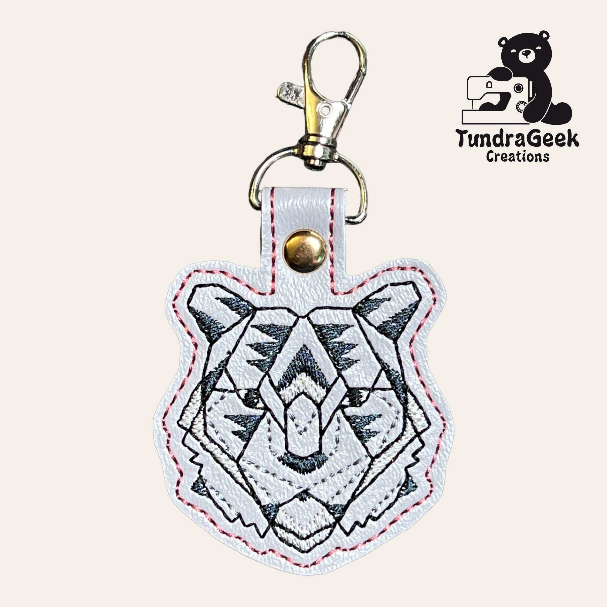 White leather keychain shaped like a tigers head with black geometric embroidery. Gold clasp attached. TundraGeek Creations logo with a bear and a sewing machine in the corner.