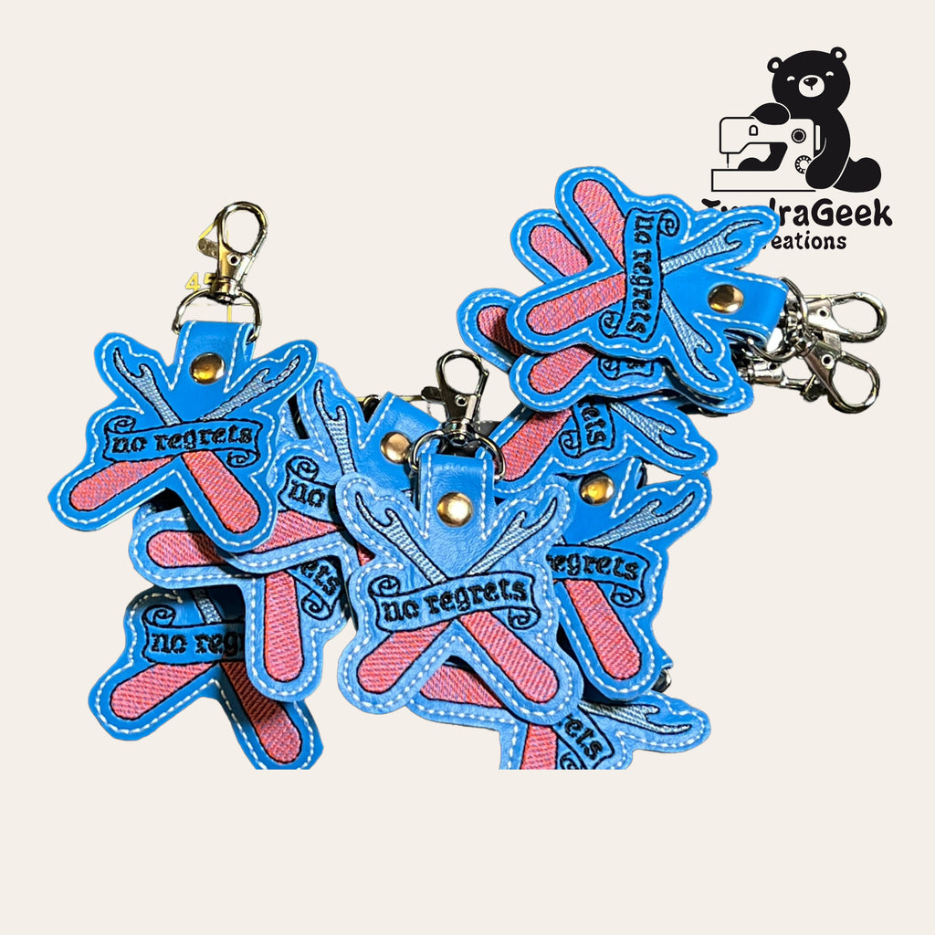A pile of blue and pink keychains shaped like crossed fingers with the words no regrets, featuring metal clasps. A bear with a sewing machine and ParaGeek Creations logo is in the corner.