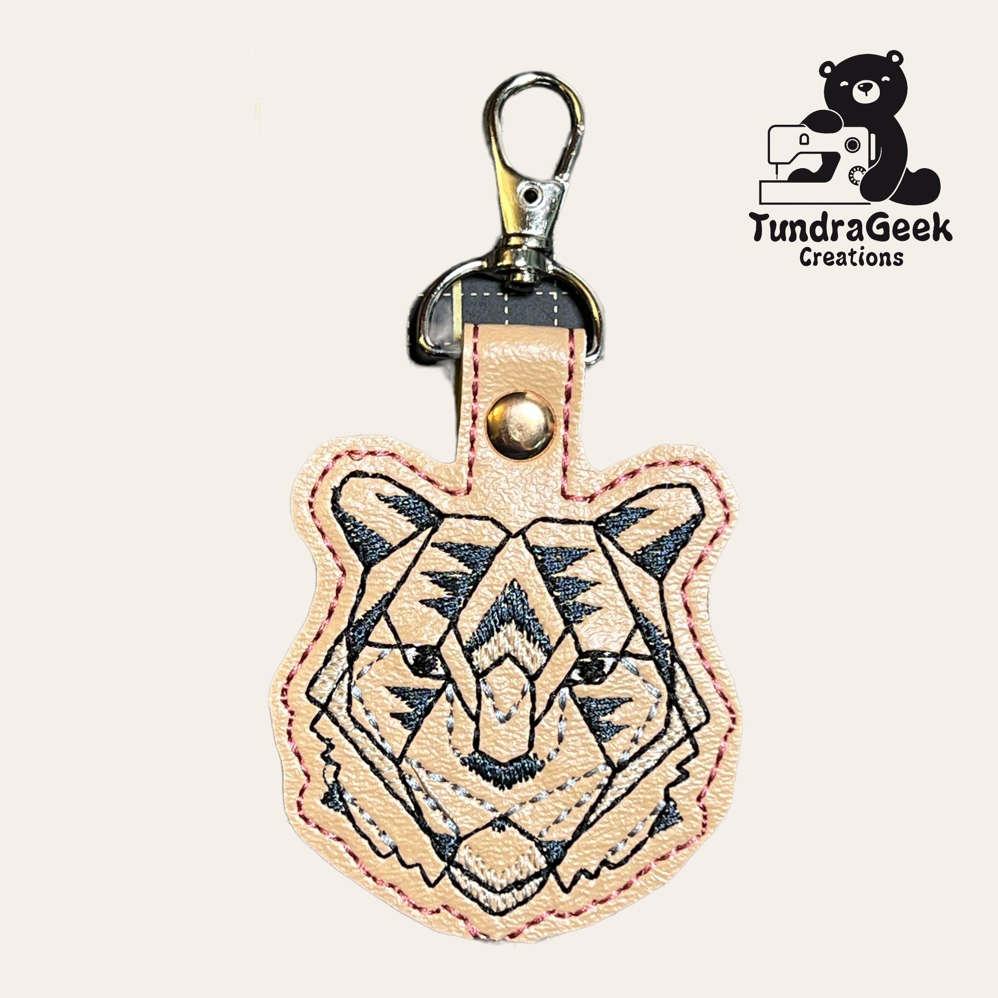 Tan leather keychain shaped like a tigers face with black embroidered details and a metal clasp; TundraGeek Creations logo is visible in the top right corner.