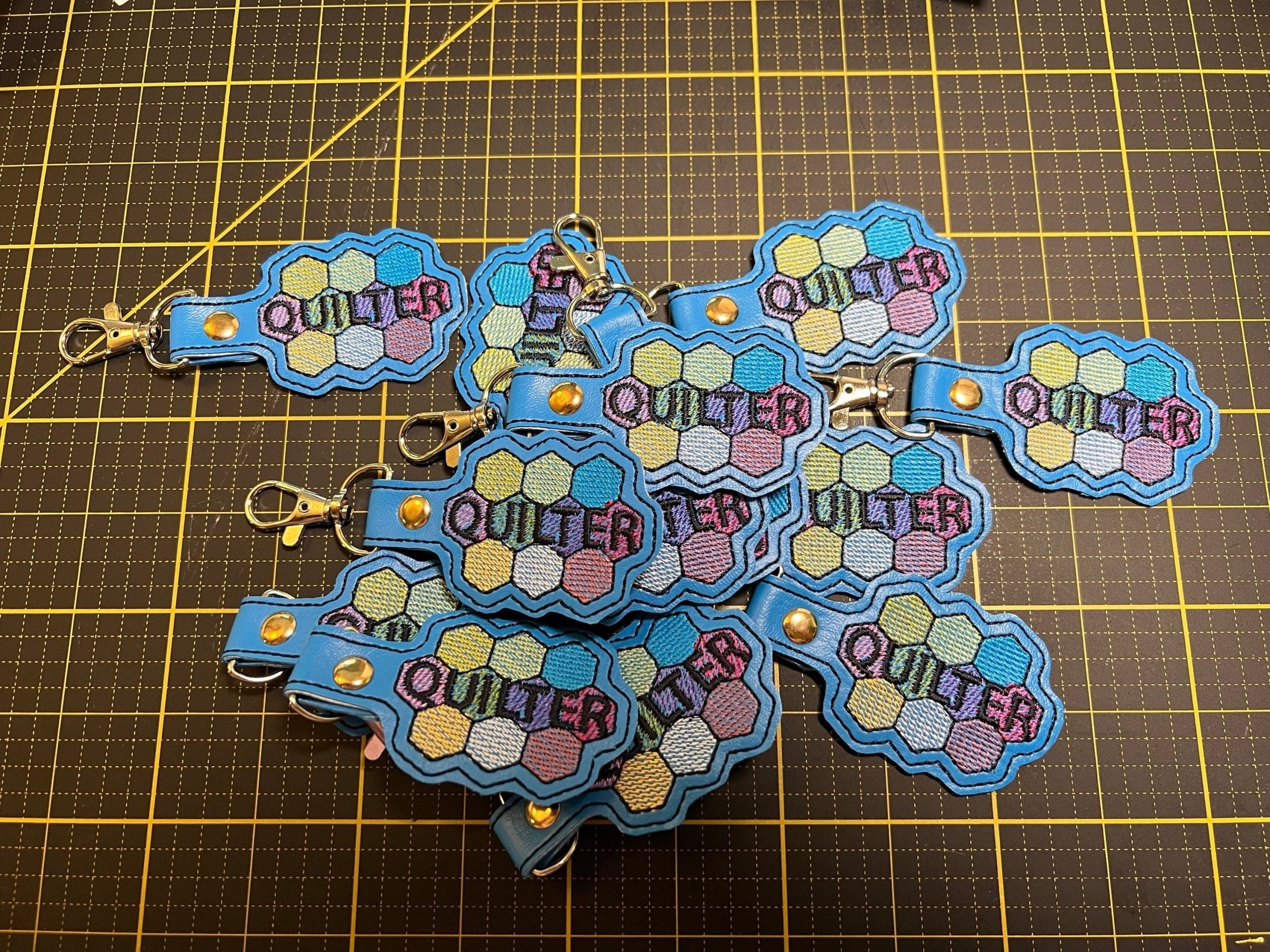 A pile of keychains with a blue border, hexagon pattern, and the word QUILTER stitched in purple, lying on a yellow grid cutting mat.