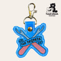 Blue and pink keychain with embroidered crossed crochet hooks and the phrase No regrets, attached to a metal clasp. TundraGeek Creations logo with bear and sewing machine in top right.