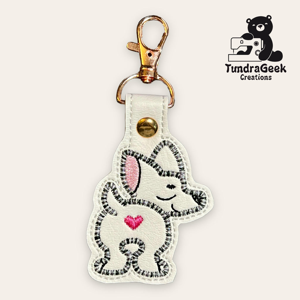 Embroidered keychain with a cartoon dog, pink ear and heart, on white faux leather with a metal clasp. Logo in the top right corner reads TundraGeek Creations with a bear and sewing machine.