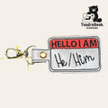 A keychain shaped like a Hello I Am name tag with He/Him written on it, featuring a metal clasp and the TundraGeek Creations logo in the corner.