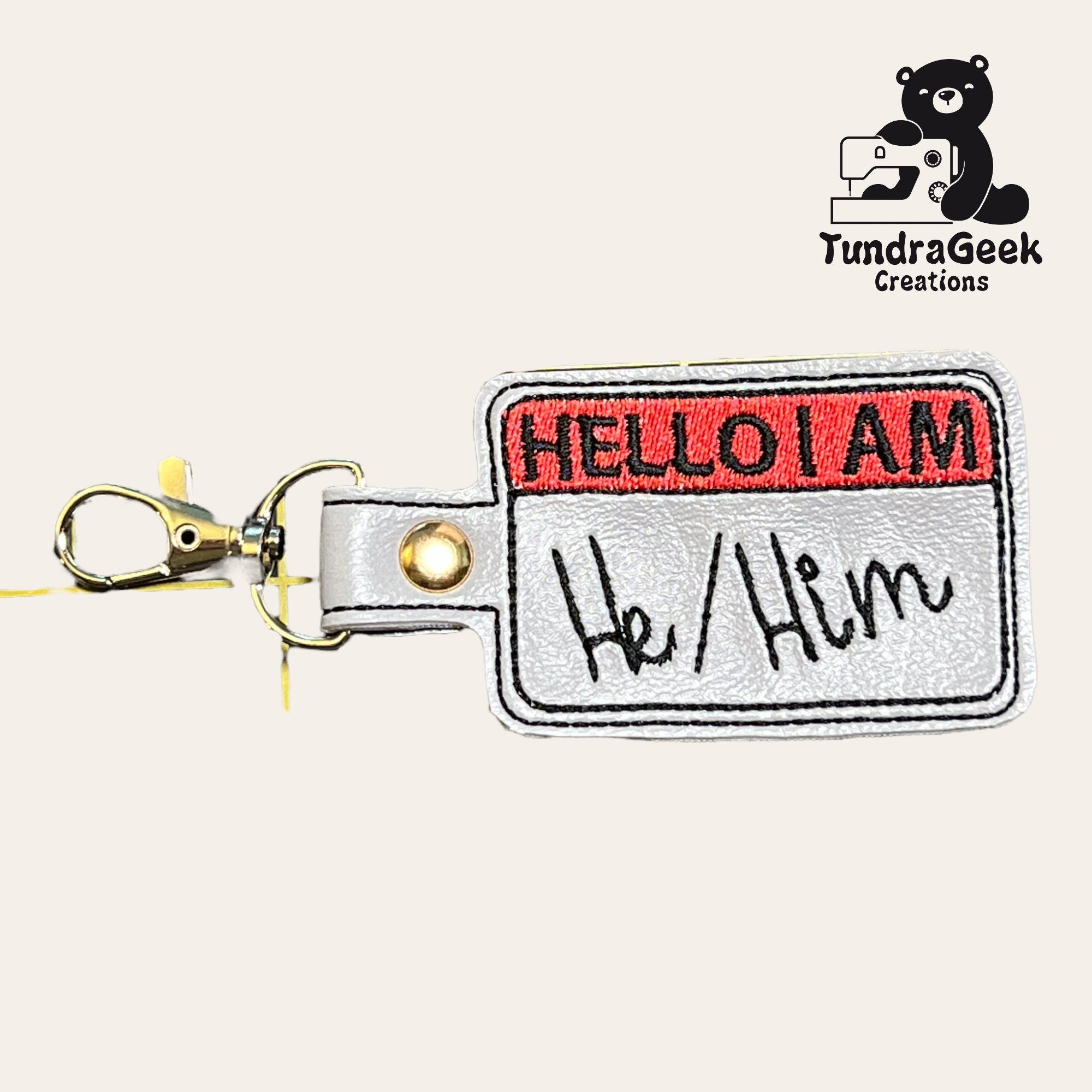A keychain shaped like a Hello I Am name tag with He/Him written on it, featuring a metal clasp and the TundraGeek Creations logo in the corner.
