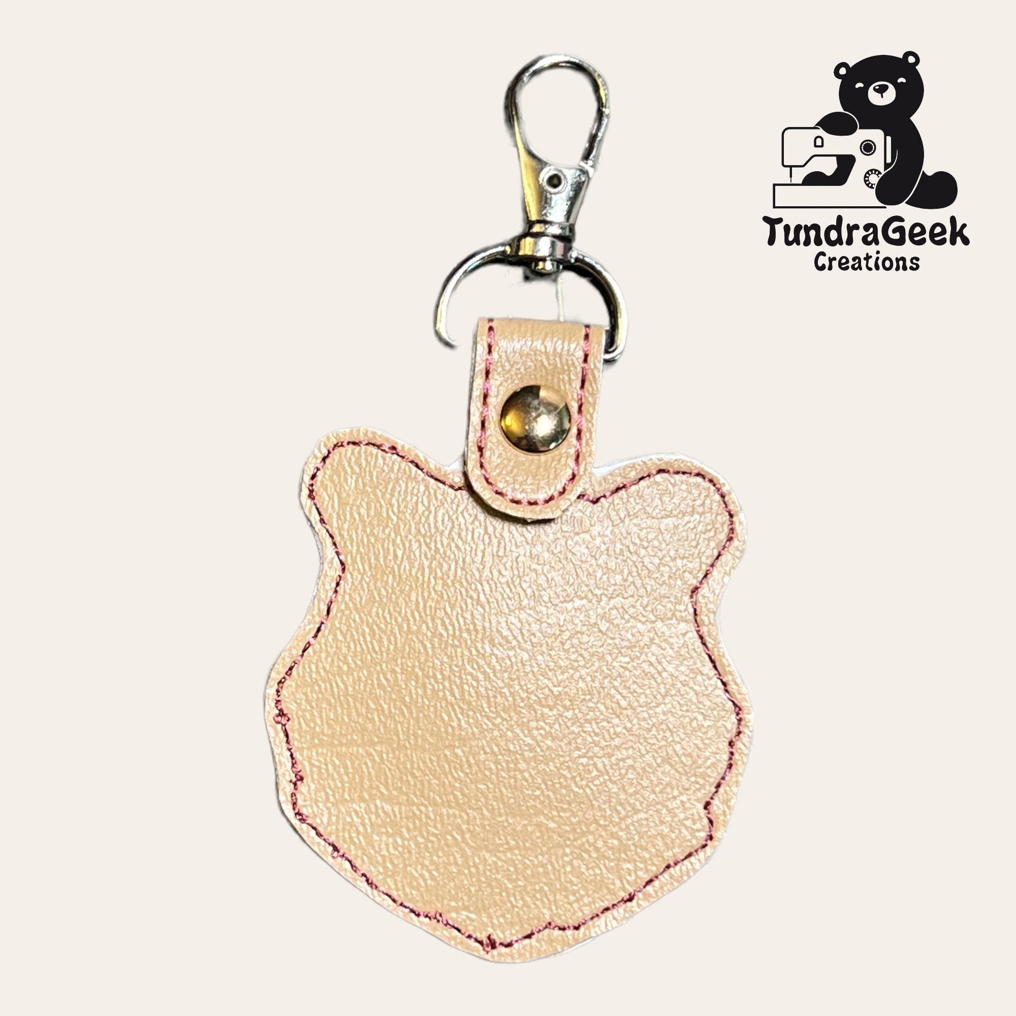 A beige, bear-shaped leather keychain with pink stitching and a metal clasp, displayed next to the TundraGeek Creations logo.