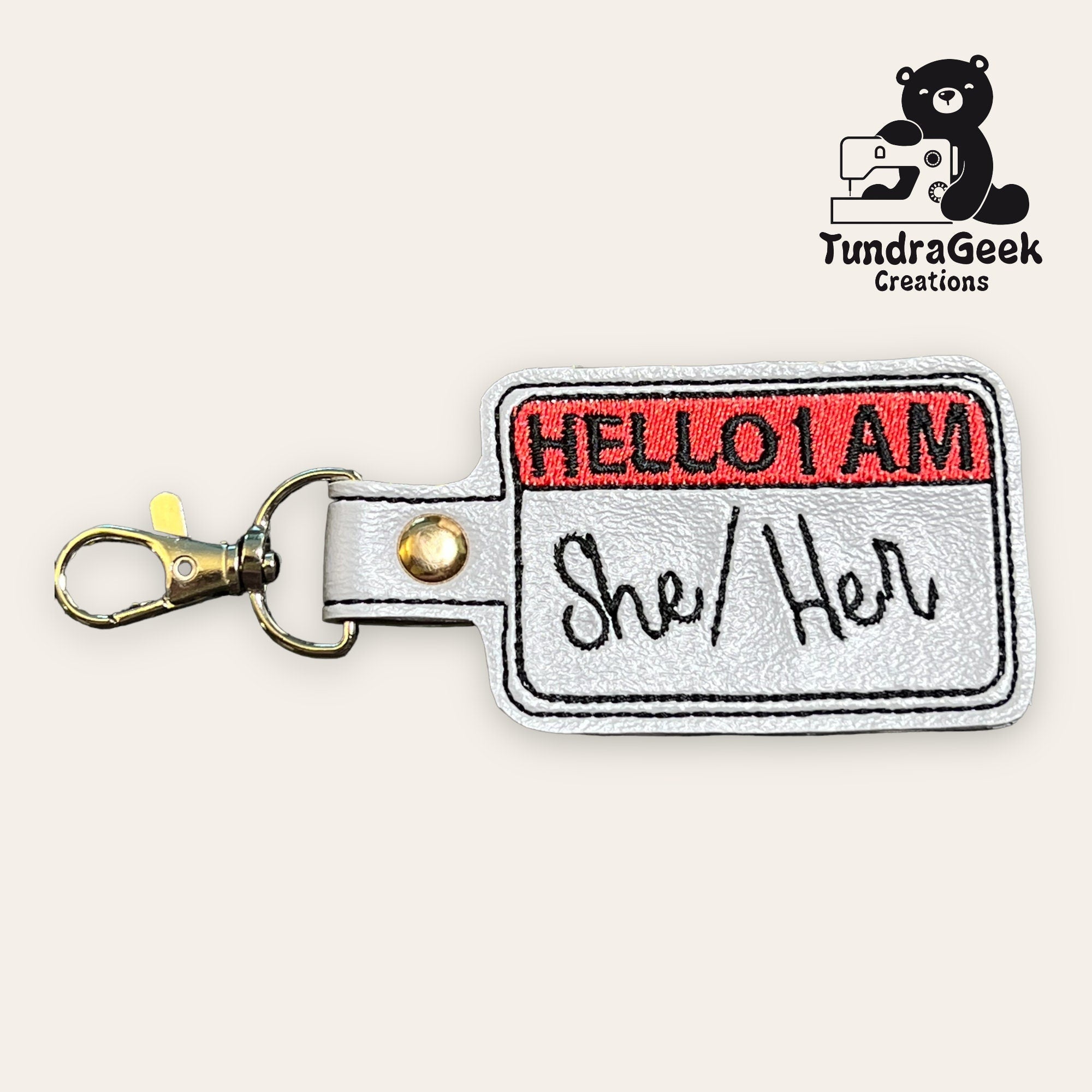 A keychain shaped like a Hello I Am name tag with She/Her written on it, and a metal clasp attached. TundraGeek Creations logo in the top right corner.