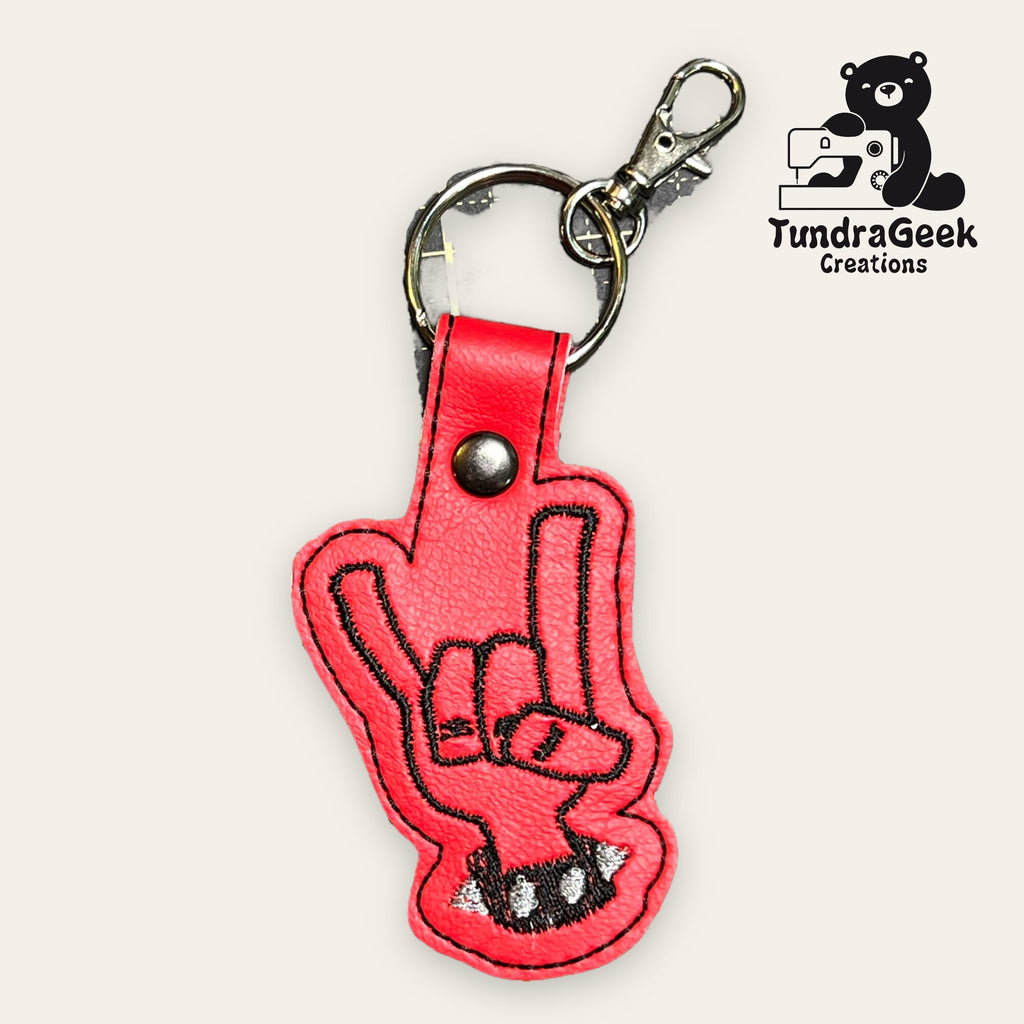 Red faux leather keychain shaped like a hand making the rock on gesture, with black embroidery and a metal clasp. TundraGeek Creations logo is in the upper right corner.