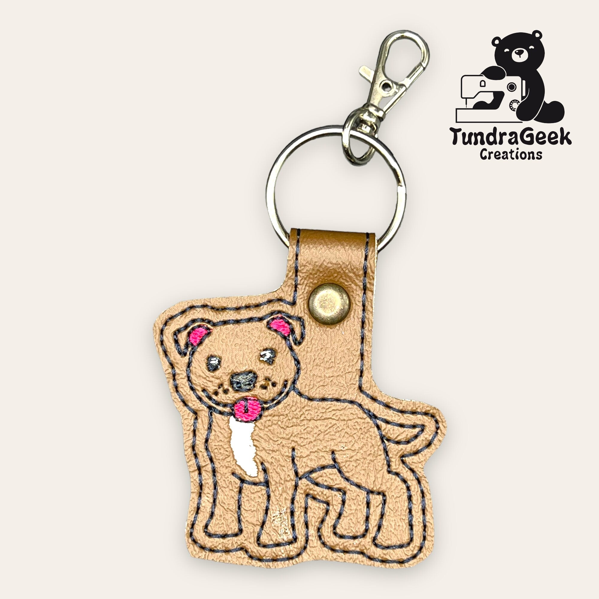 Tan faux leather dog-shaped keychain with embroidered details, a metal key ring, and a lobster clasp. TundraGeek Creations logo is in the top right corner.