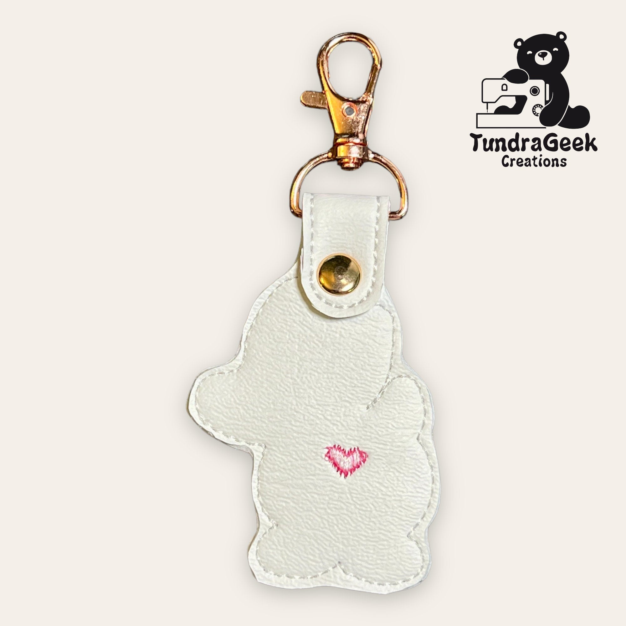 White bear-shaped keychain with pink heart detail, gold hardware, and snap button. TundraGeek Creations logo with a bear and sewing machine in the corner.